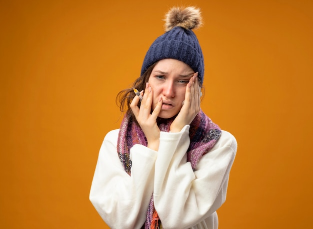 Triste giovane ragazza malata che indossa veste bianca e cappello invernale con sciarpa che tiene la siringa con le pillole che mettono le mani sulle guance isolate sull'arancia