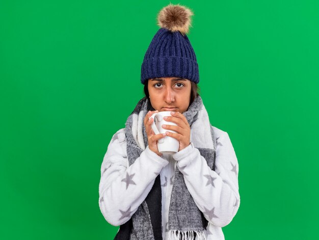 緑の背景で隔離のお茶のカップを保持しているスカーフと冬の帽子をかぶって悲しい若い病気の女の子