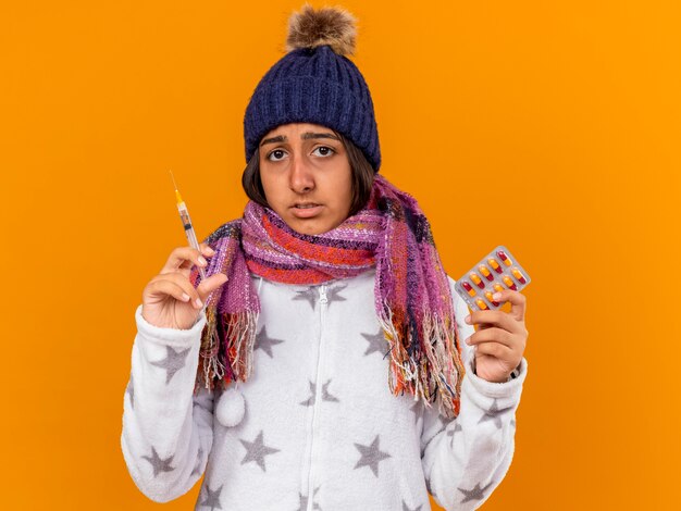 Sad young ill girl wearing winter hat with scarf holding syringe with pills isolated on yellow background