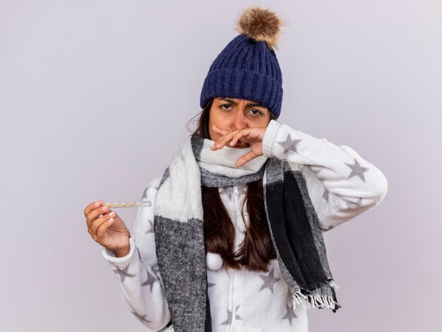 Sad young ill girl wearing winter hat with scarf holding thermometer and wiping nose with hand isolated on white background