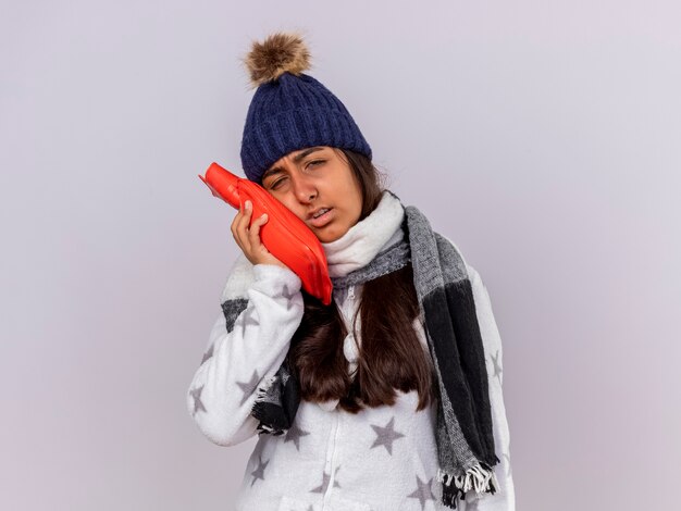 Грустная молодая больная девушка в зимней шапке с шарфом кладет мешок с горячей водой на щеку на белом фоне