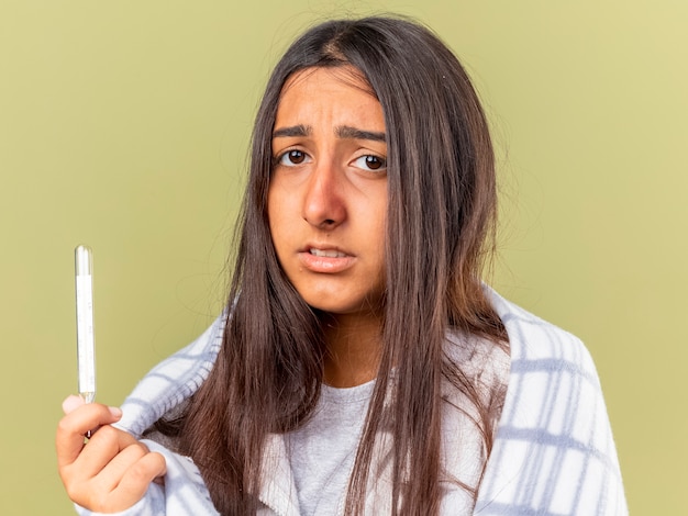 Foto gratuita triste giovane ragazza malata avvolta in plaid tenendo il termometro isolato su verde oliva