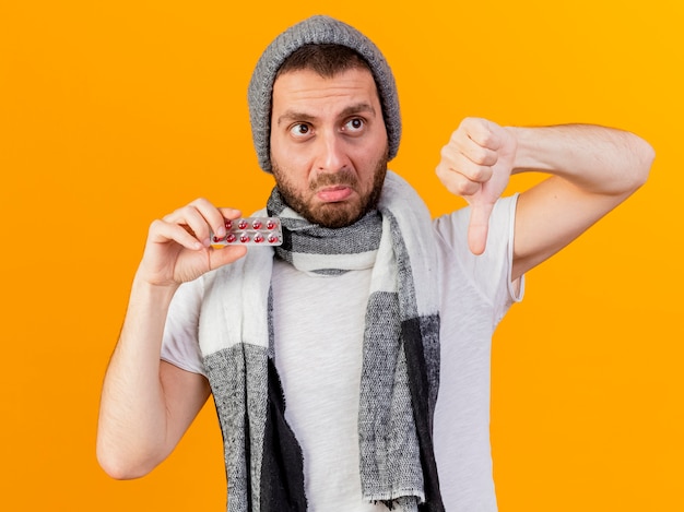 Sad young ill man wearing winter hat and scarf holding pills and showing thumb down isolated on yellow background