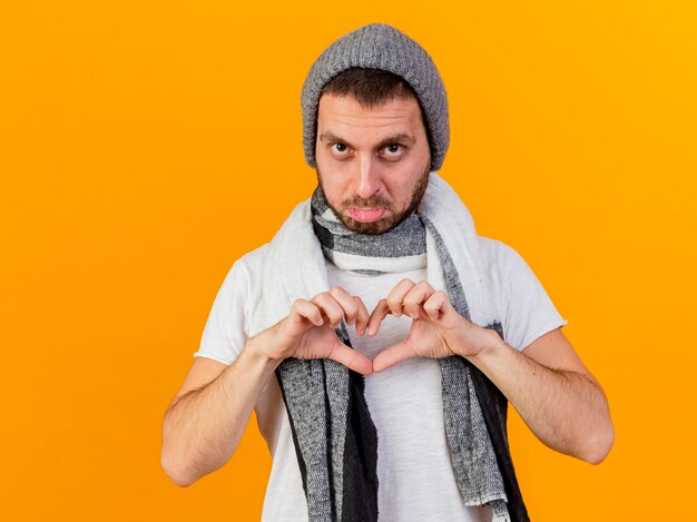 Sad young ill man wearing winter hat and scarf showing heart gesture isolated on yellow background