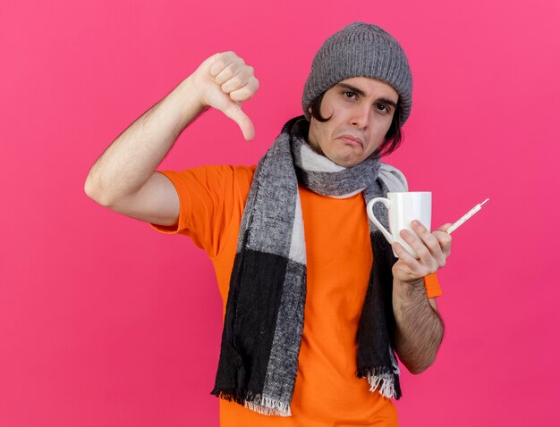 Sad young ill man wearing winter hat with scarf holding cup of tea with thermometer showing thumb down isolated on pink background