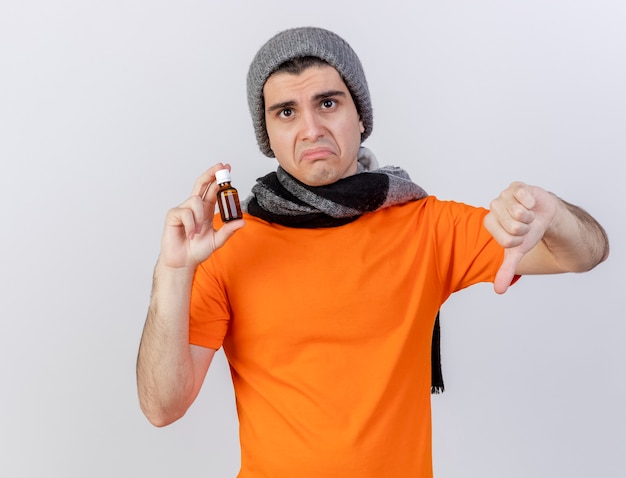 Sad young ill man wearing winter hat with scarf holding medicine in glass bottle showing thumb down