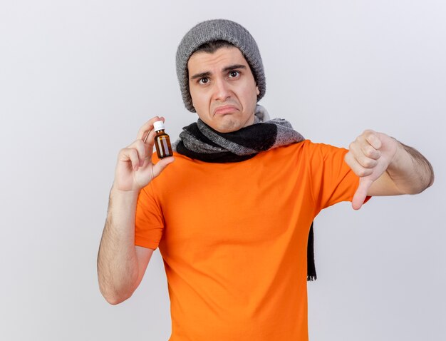 Sad young ill man wearing winter hat with scarf holding medicine in glass bottle showing thumb down