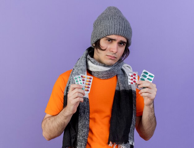 sad young ill man wearing winter hat with scarf holding pills isolated on purple