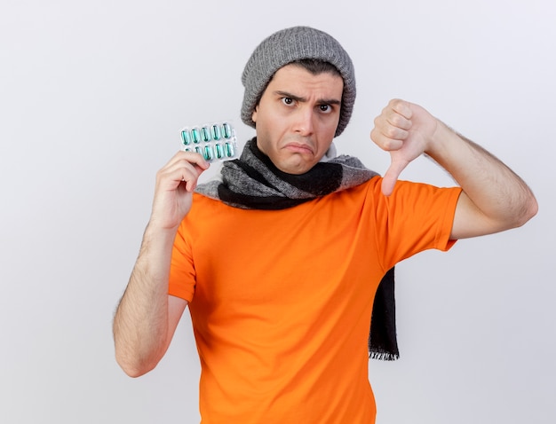 Грустный молодой больной человек в зимней шапке с шарфом держит таблетки, показывая большой палец вниз на белом фоне