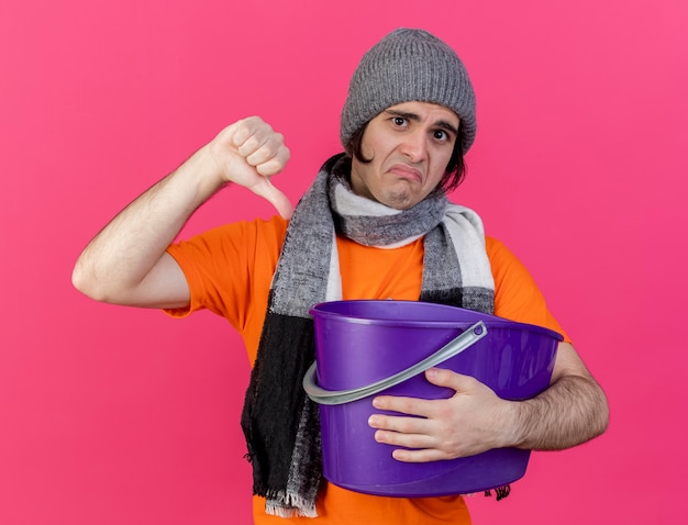 Грустный молодой больной человек в зимней шапке с шарфом держит пластиковое ведро, показывая большой палец вниз, изолированный на розовом