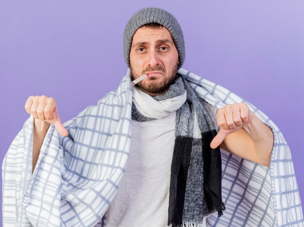 Sad young ill man wearing winter hat with scarf putting thermometer in mouth wrapped in plaid and showing thumbs down isolated on purple