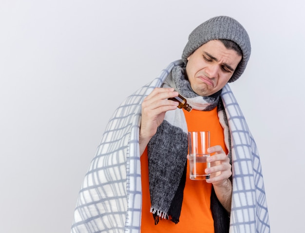 Triste giovane uomo malato che indossa un cappello invernale con sciarpa avvolta in plaid versando medicamento in un bicchiere d'acqua