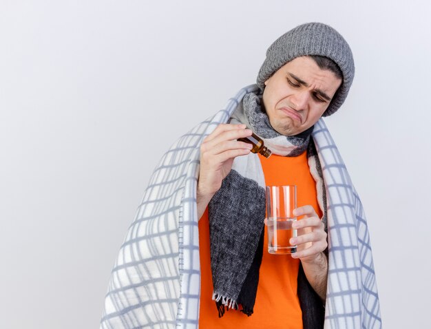 Sad young ill man wearing winter hat with scarf wrapped in plaid pouring medicament into glass of water