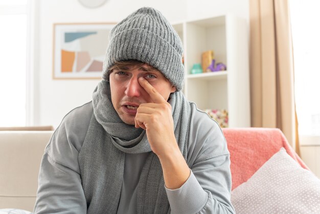 sad young ill man with scarf around neck wearing winter hat putting finger on his eyelid sitting on couch at living room