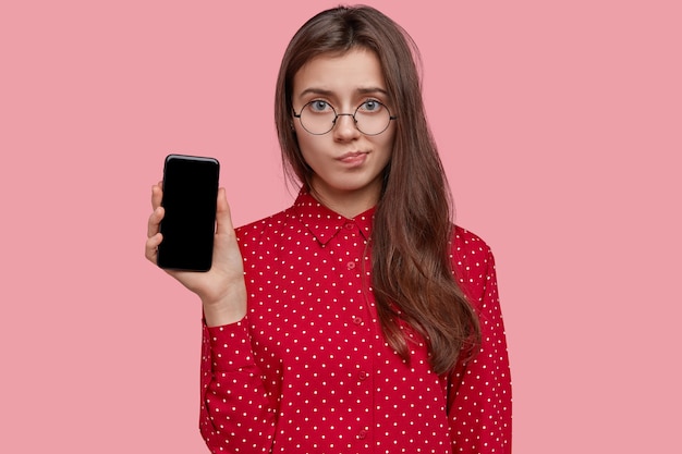 Sad young lady purses lips, holds modern cell phone with mockup screen, wears transparent glasses, dressed in polka dot shirt