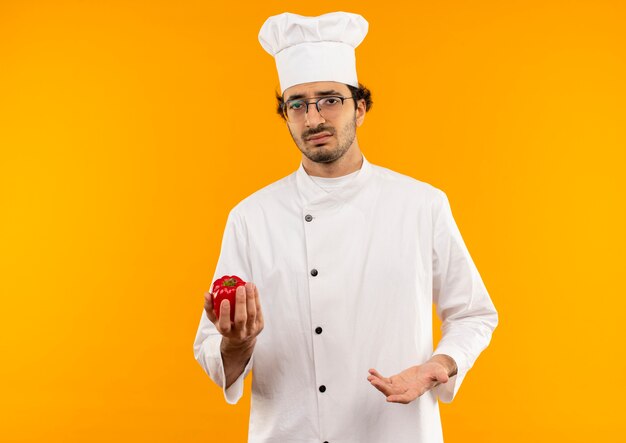 Sad young male cook wearing chef uniform and glasses holding pepper isolated on yellow wall