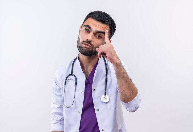Грустный молодой мужчина-врач в медицинском халате со стетоскопом положил палец на лоб на изолированном белом фоне