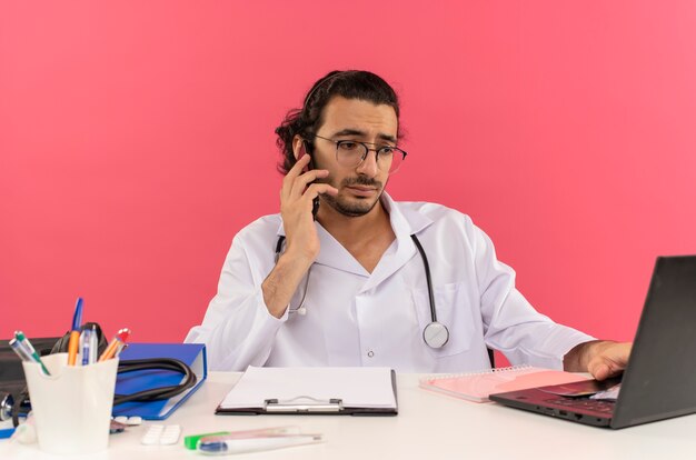 Sad young male doctor with medical glasses wearing medical robe with stethoscope sitting at desk