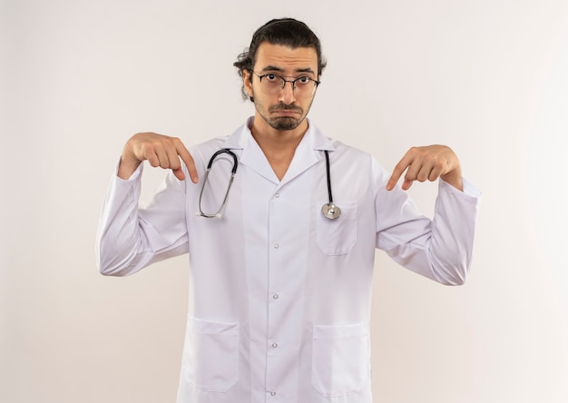 Free photo sad young male doctor with optical glasses wearing white robe with stethoscope points to down on isolated white wall with copy space