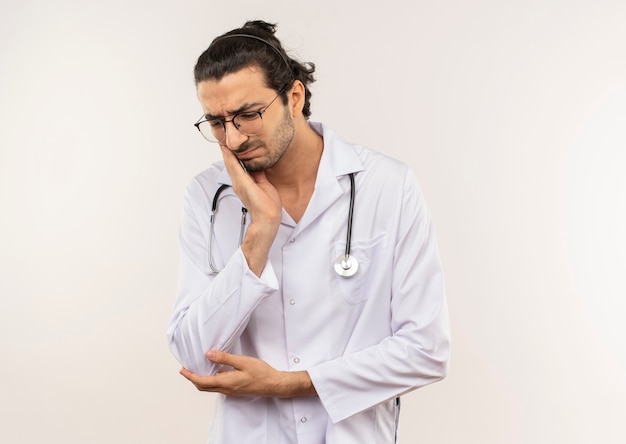 Sad young male doctor with optical glasses wearing white robe with stethoscope putting hand on aching tooth on isolated white wall with copy space