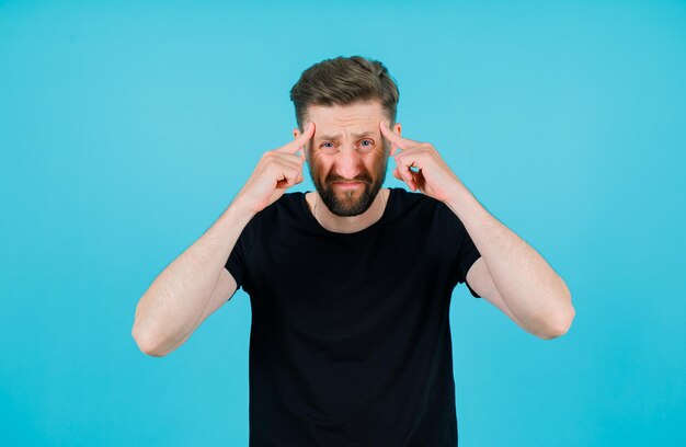 Sad young man is holding forefingers on temples on blue background