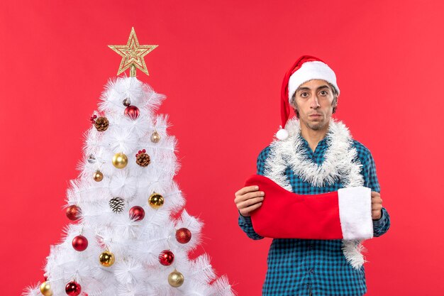 Грустный молодой человек в шляпе санта-клауса в синей полосатой рубашке и держит рождественский носок возле рождественской елки на красном