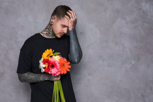 Sad young man with tattoo on his body holding fresh colorful gerbera flowers