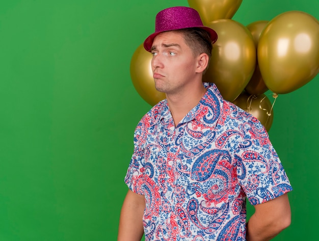 Sad young party guy looking at side wearing pink hat standing infront of balloons isolated on green