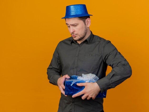 sad young party guy wearing blue hat holding gift box isolated on orange