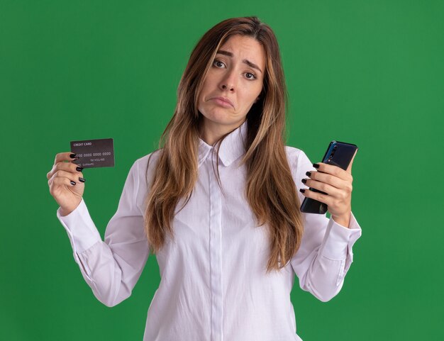 Sad young pretty caucasian girl holds credit card and phone isolated on green wall with copy space