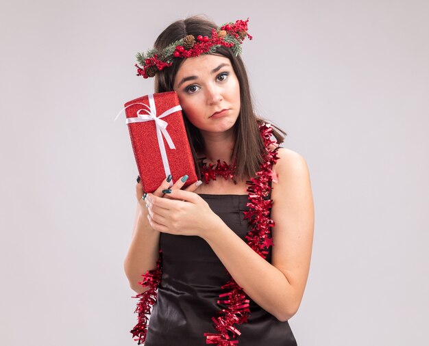 Sad young pretty caucasian girl wearing christmas head wreath and tinsel garland around neck holding gift package touching face with it looking at camera isolated on white background with copy space