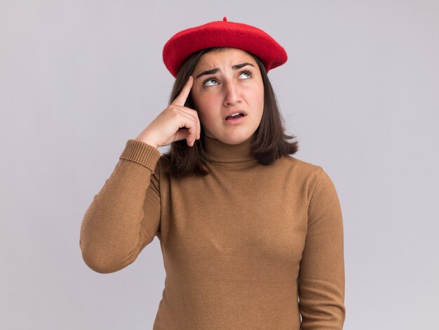 Sad young pretty caucasian girl with beret hat puts finger on temple and looks up