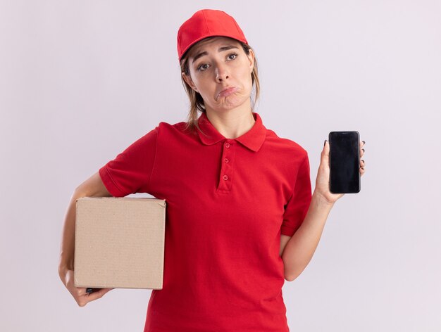 Sad young pretty delivery woman in uniform holds cardbox and phone isolated on white wall