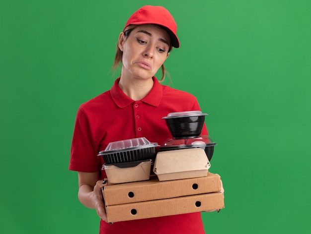 La giovane donna graziosa di consegna triste in uniforme tiene i pacchetti e i contenitori di cibo di carta sulle scatole della pizza isolate sulla parete verde