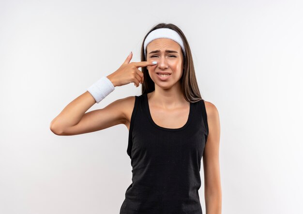 Грустная молодая симпатичная спортивная девушка с головной повязкой и браслетом, указывающая на нос на белом пространстве