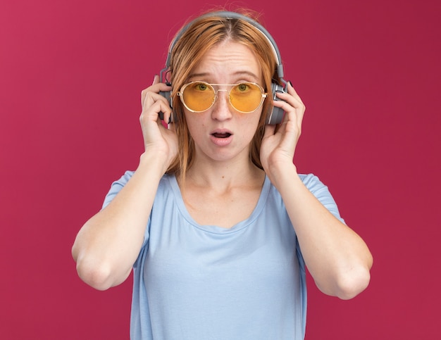 Sad young redhead ginger girl with freckles in sun glasses and on headphones  isolated on pink wall with copy space