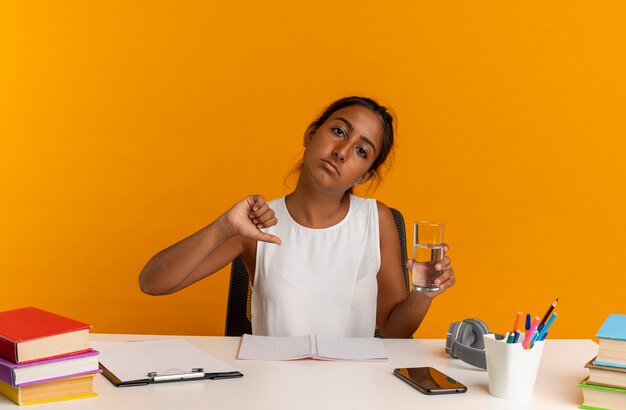 Sad young schoolgirl sitting at desk with school tools holding glass of water her thumb down isolated on orange wall
