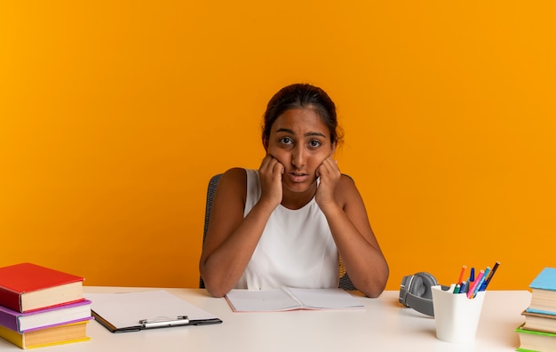 Sad young schoolgirl sitting at desk with school tools putting hands on cheeks isolated on orange wall