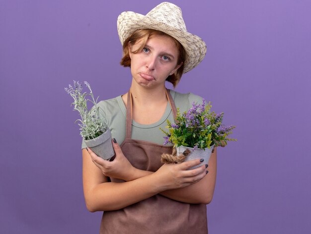紫色の植木鉢に花を保持しているガーデニング帽子をかぶっている悲しい若いスラブ女性の庭師