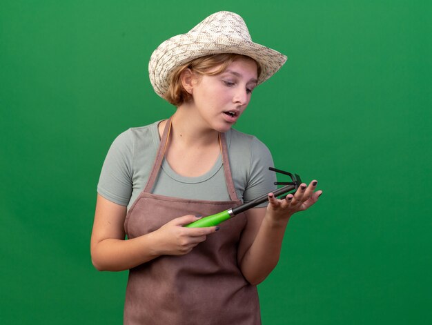 Sad young slavic female gardener wearing gardening hat holding and looking at hoe rake on green