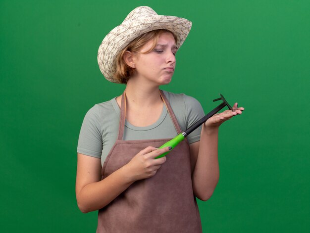 Sad young slavic female gardener wearing gardening hat holds and looks at hoe rake on green