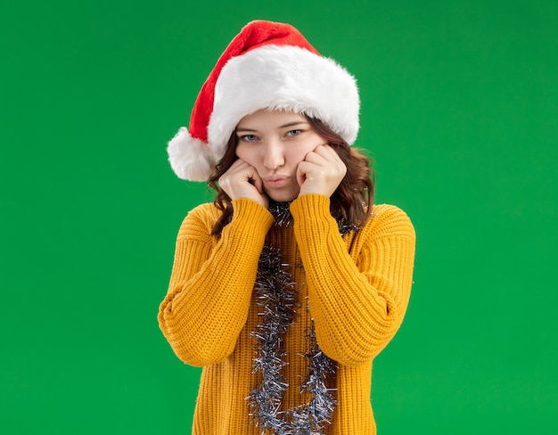 sad young slavic girl with santa hat and with garland around neck puts hands on face isolated on green background with copy space