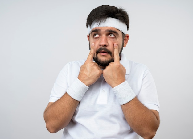 Sad young sporty man looking at side wearing headband and wristband pulling down eye lids isolated on white wall