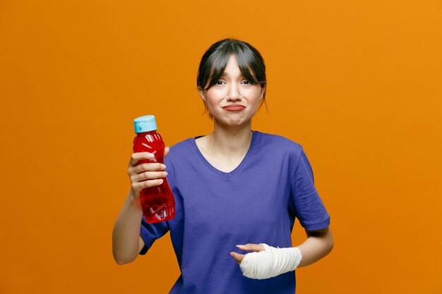 オレンジ色の背景に分離された包帯で包まれた手首と水のボトルを示すカメラを見て手を空中に保つTシャツを着て悲しい若いスポーティな女性