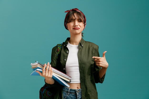Sad young student girl wearing bandana and backpack holding note pads with pen pointing at them looking at camera isolated on blue background