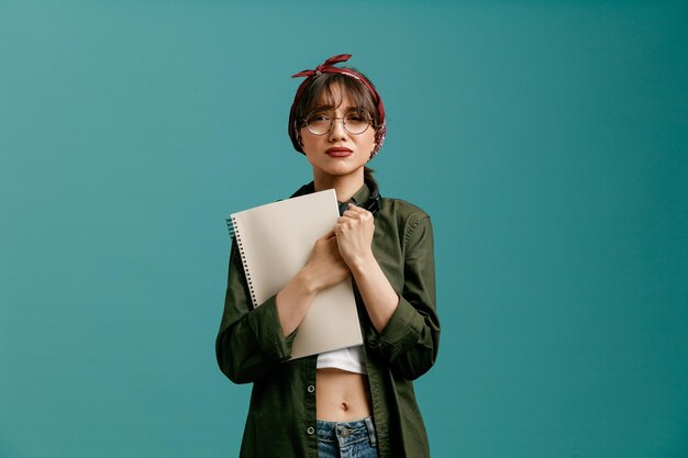 Sad young student girl wearing bandana glasses and headphones around neck holding large note pad hugging it keeping hands together looking at camera isolated on blue background