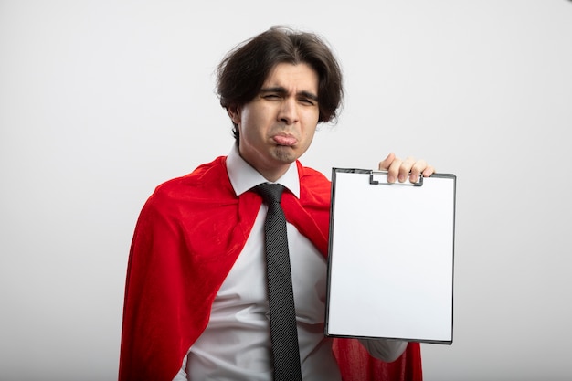 Sad young superhero guy wearing tie holding clipboard isolated on white
