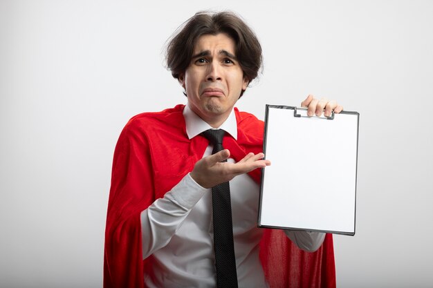 Sad young superhero guy wearing tie holding and points with hand at clipboard isolated on white