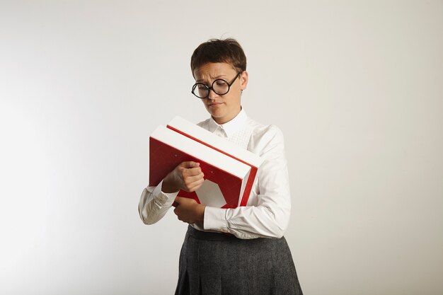 Sad young teacher in white blouse and gray tweed skirt looks at thick and heavy binders with sadness isolated on white