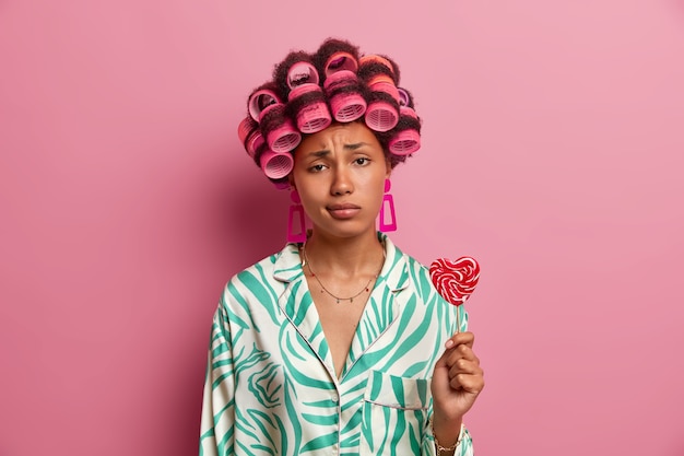 Free photo sad young woman gets hair permed, looks unhappily, wears hair rollers, dressed in casual clothes, holds sweet lollipop, spends time on herself, caring about her beauty, isolated on pink wall